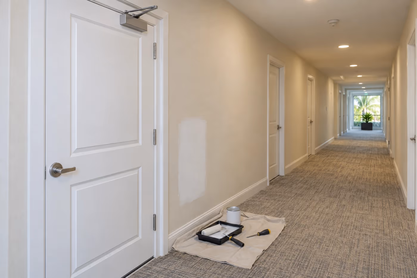 Condominium common hallway with white unit doors, a door closer on the nearest door, and a primed wall patch above a drop cloth with a paint tray and roller