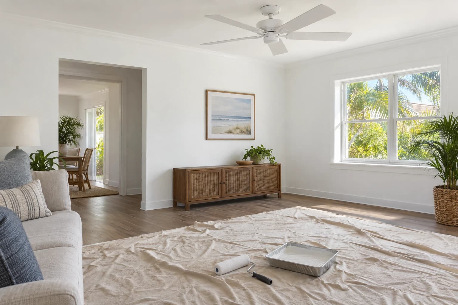A freshly painted living room in soft coastal white with a new white ceiling fan, a paint roller and tray resting on a canvas drop cloth