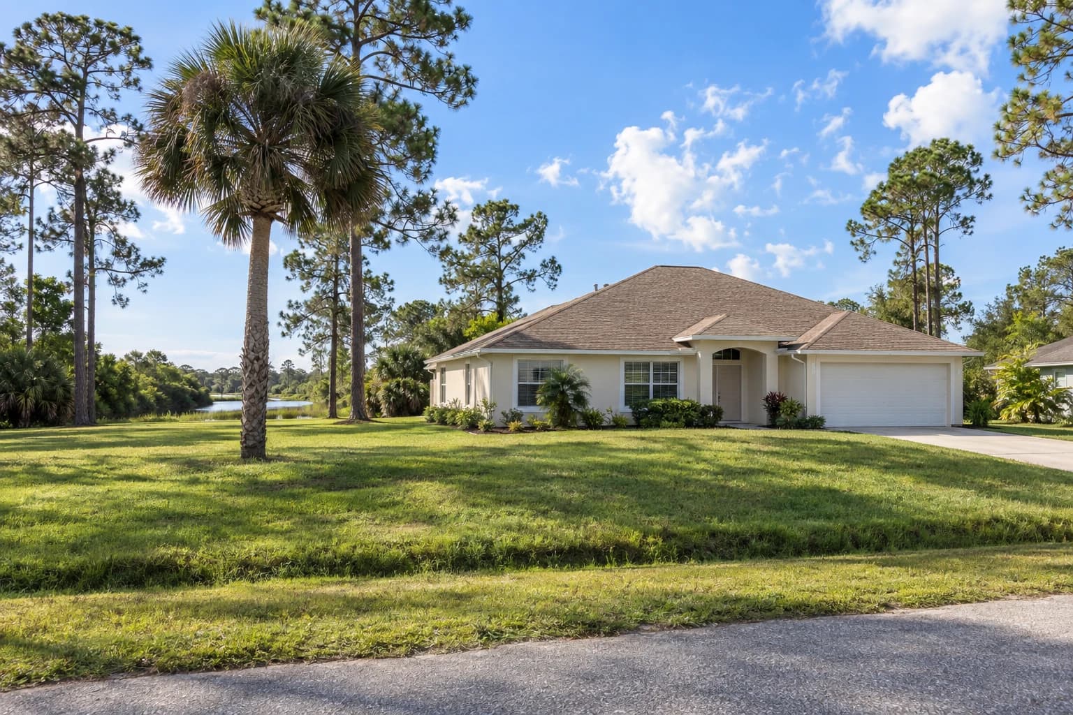 One-story concrete block home with cream stucco, a hip shingle roof, and a wide grass lot with slash pines and a cabbage palm on a quiet North Port street