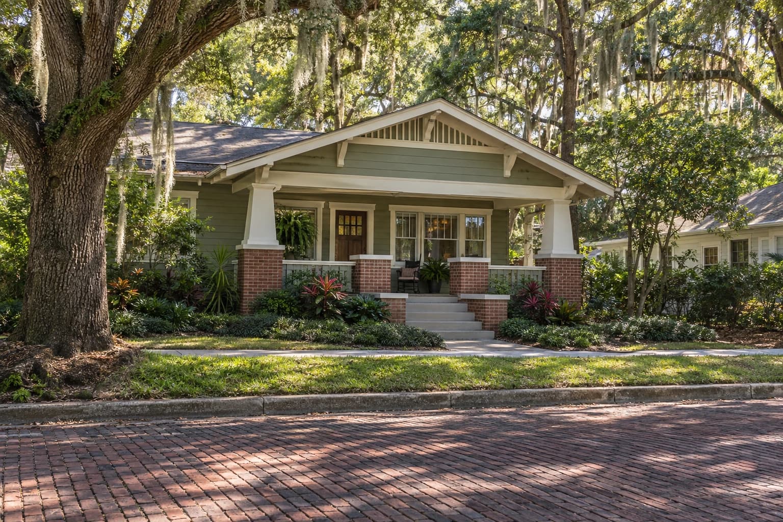 A 1920s Craftsman bungalow with a deep front porch and tapered porch columns on a brick street, shaded by live oaks