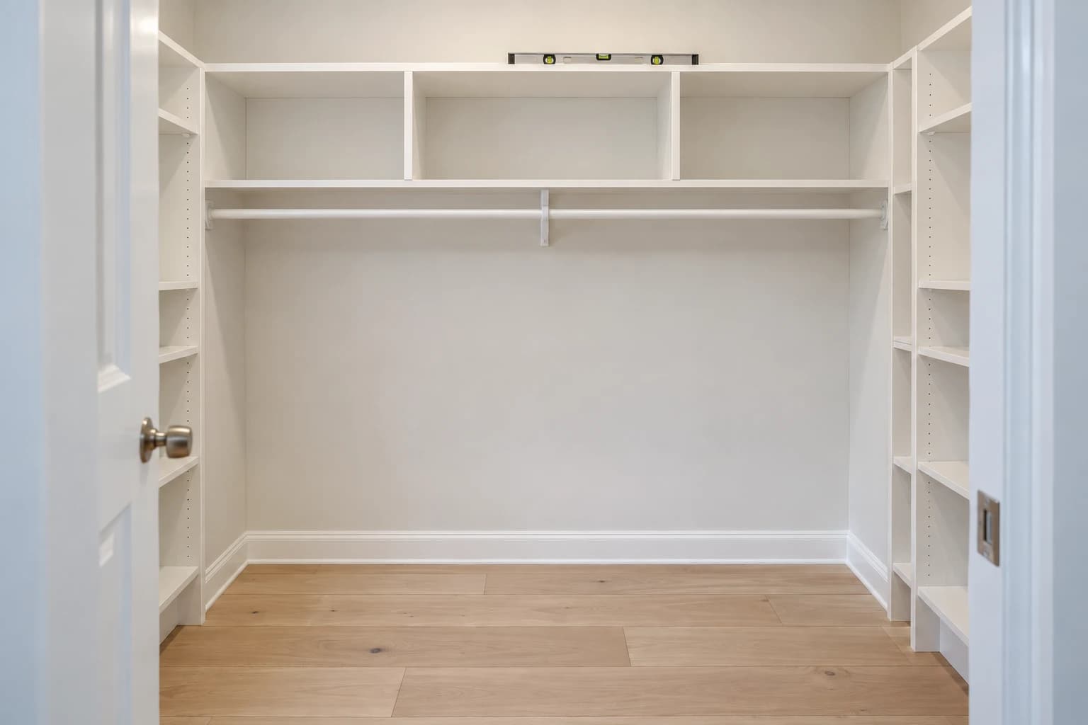 Walk-in closet with new white wood shelving and a hanging rod built wall to wall, with a level resting on the top shelf