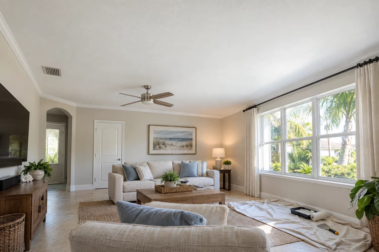 A freshly patched and painted flat white ceiling over a Florida living room, with a drywall knife and a small roller tray resting on a drop cloth by the window