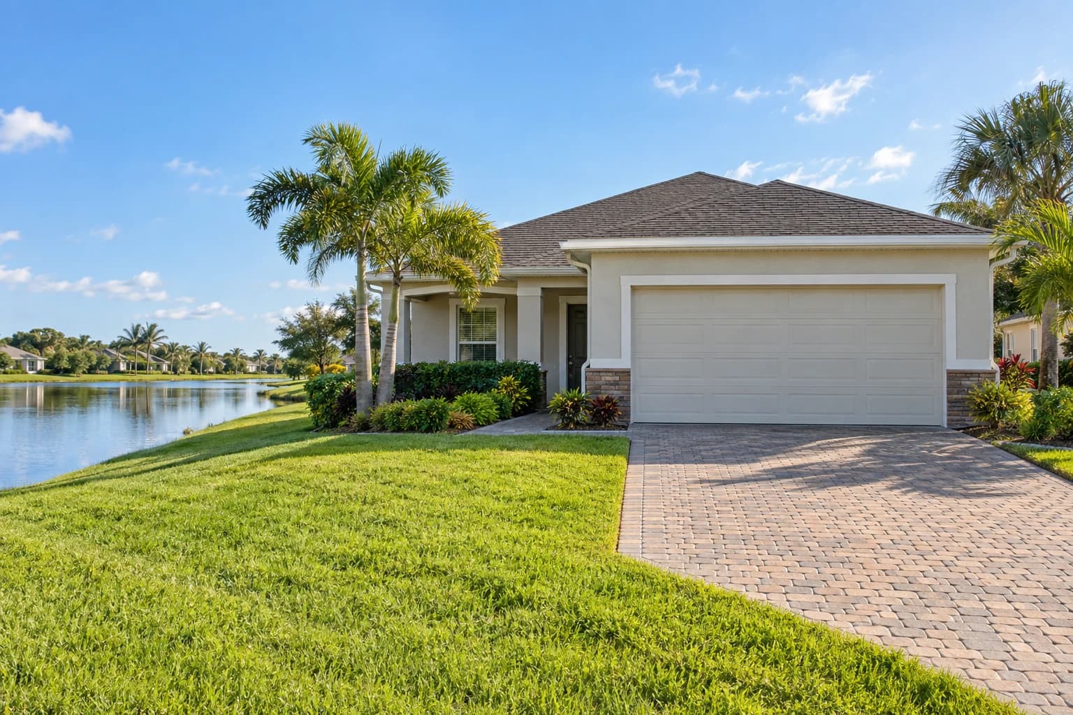Newer one-story stucco home with a tile-look roof, a two-car garage, and palms beside a retention pond in a West Melbourne subdivision