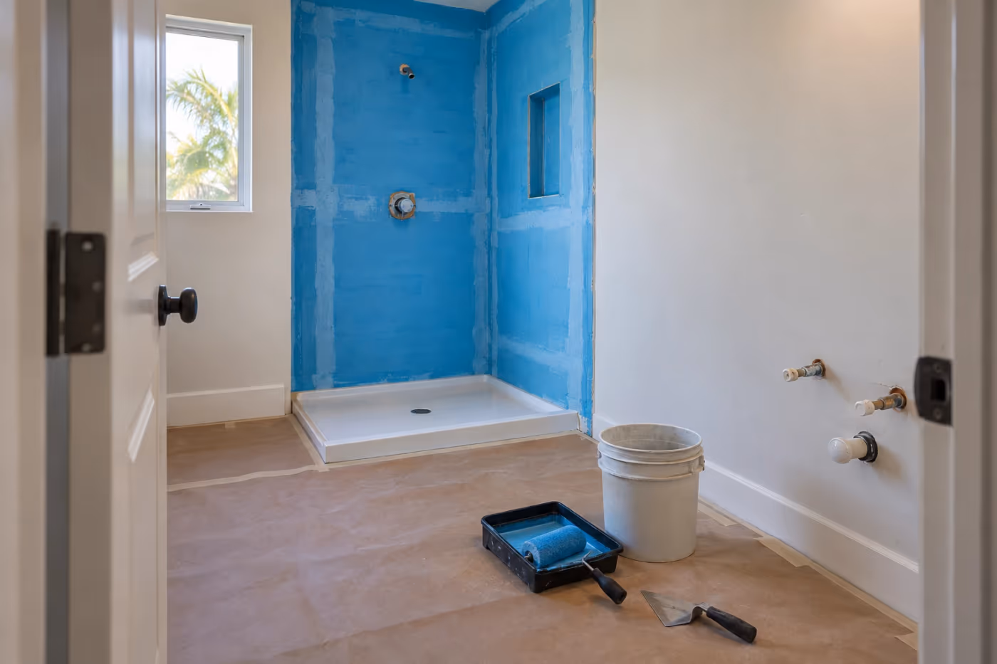 A Florida hall bathroom mid remodel, the shower walls covered in backer board with blue waterproofing rolled on, a new shower pan in place, capped water lines where the vanity will go, and a roller tray and bucket on protective floor paper