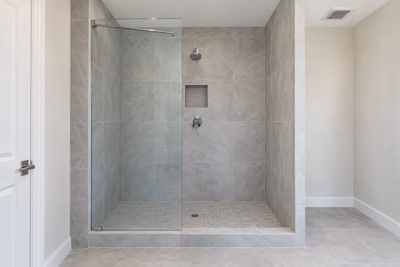 A finished tiled walk-in shower with light gray porcelain walls to the ceiling, a recessed niche, a small tile floor sloping to a center drain, and one fixed frameless glass panel beside an open entry