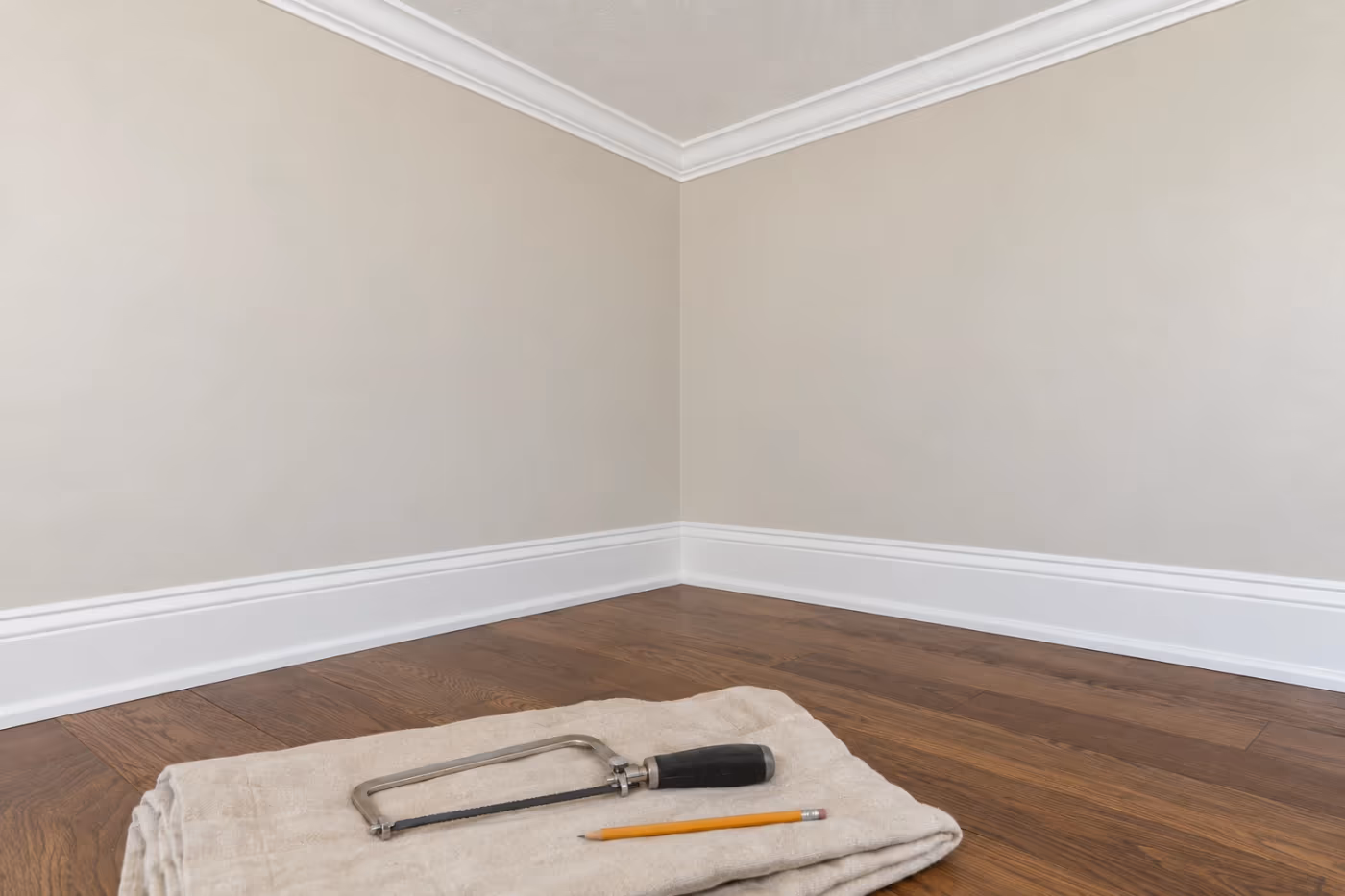 The corner of an empty Florida room where new white crown molding meets in a tight inside corner, with matching tall baseboard below and a coping saw and pencil on a folded drop cloth
