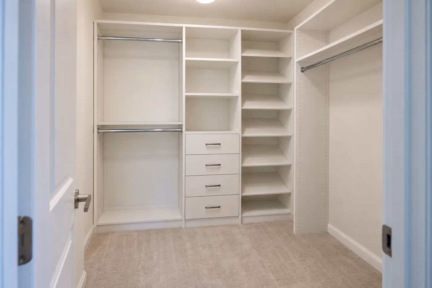 A small walk-in closet with a newly installed white closet system: double hanging rods, a long rod with a shelf above, a stack of four drawers, and angled shoe shelves, all empty and level