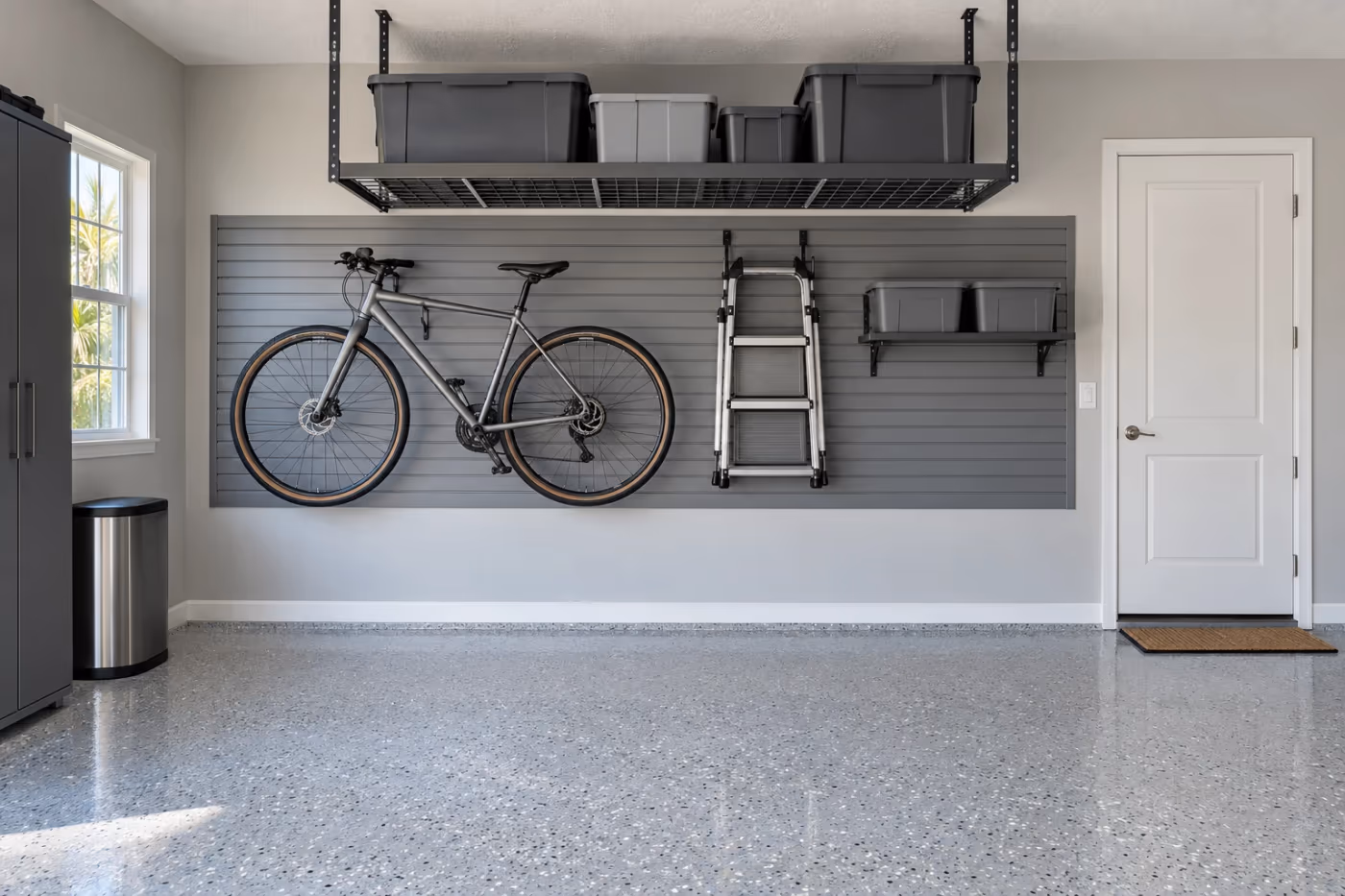 The back wall of a clean garage with a gray flake epoxy floor, gray slat wall panels holding a bicycle, a step ladder, and a shelf of gray bins, and an overhead storage rack above