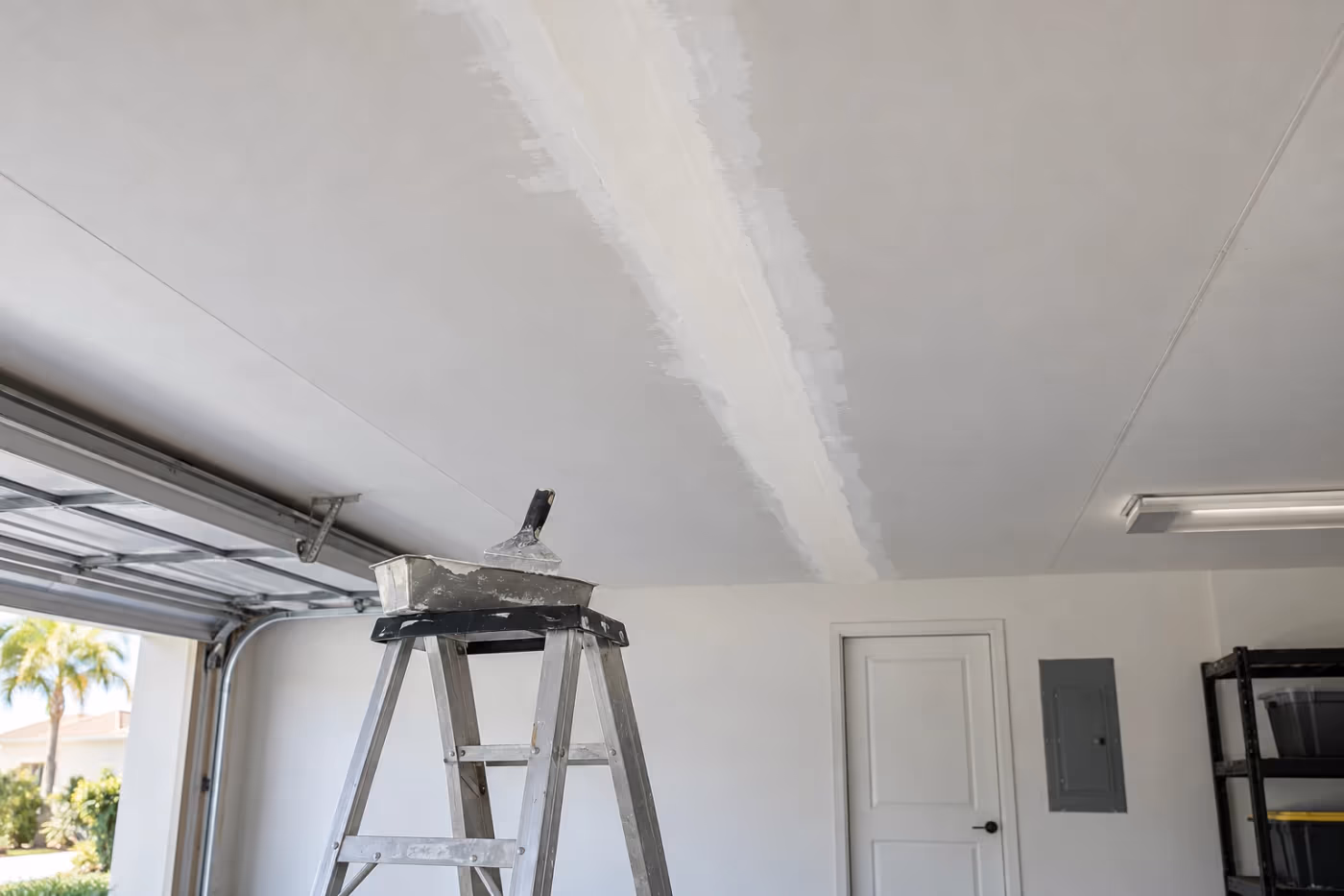 A white drywall garage ceiling with one long seam freshly retaped and feathered with off-white joint compound, an aluminum step ladder below with a mud pan and taping knife on its top