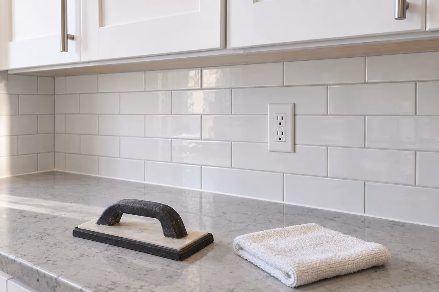 A newly installed white subway tile backsplash between a light gray quartz countertop and white upper cabinets, with an outlet cut neatly into the tile and a grout float and folded towel resting on the counter