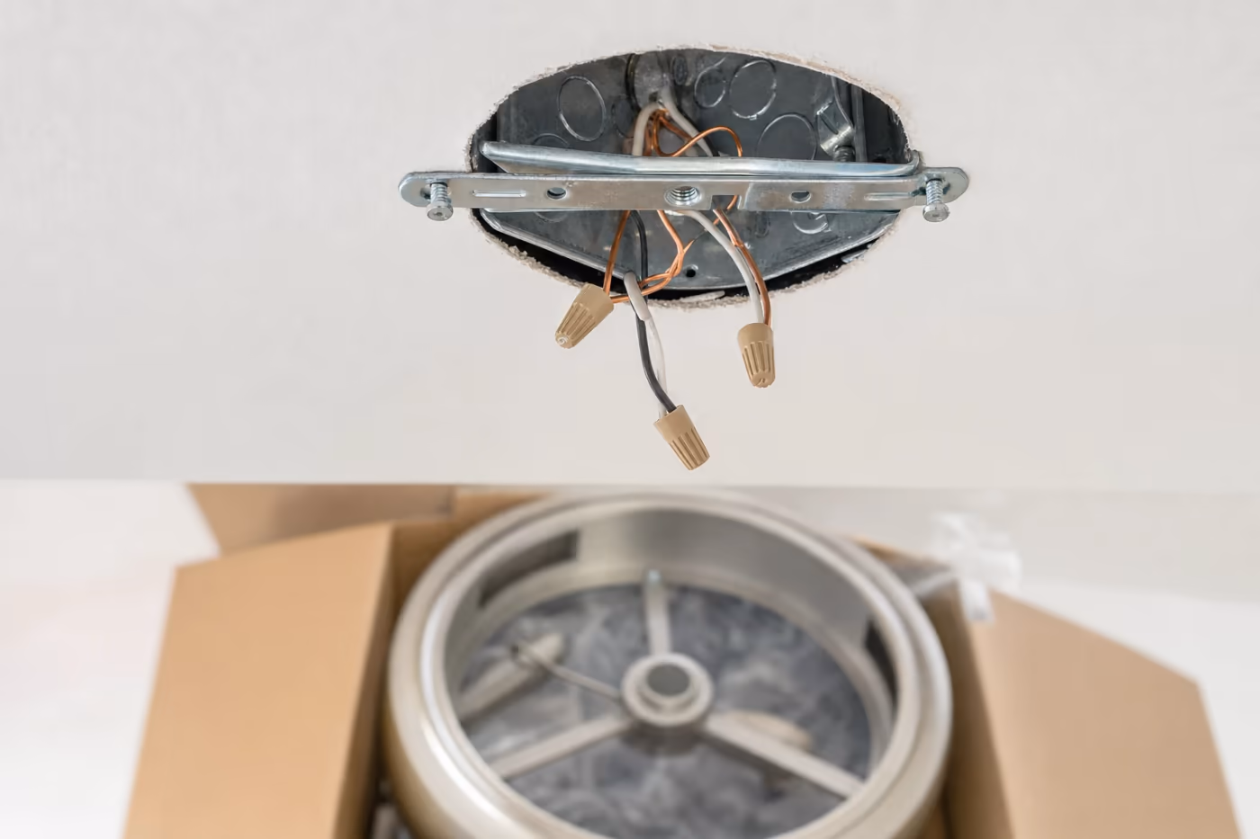 Close-up of an open ceiling junction box with mounting bracket and capped copper wires ready for a new brushed-metal light fixture installation in a Florida home