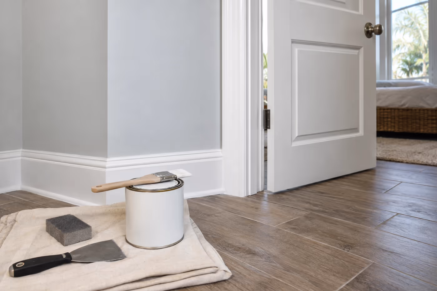 A freshly painted white door casing and tall baseboard with a clean caulk line against a pale gray wall, a brush resting on a closed quart can with a putty knife and sanding sponge on a drop cloth