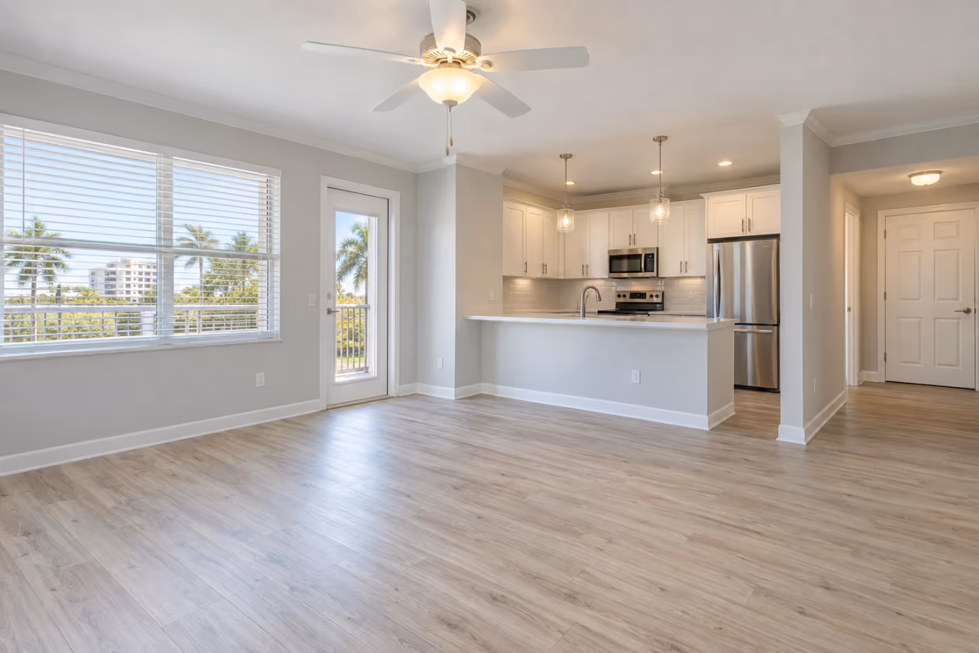 A clean, empty Florida condo living room and kitchen ready for new tenants, with light gray walls, wood-look plank floors, white cabinets, and a ceiling fan with its light on