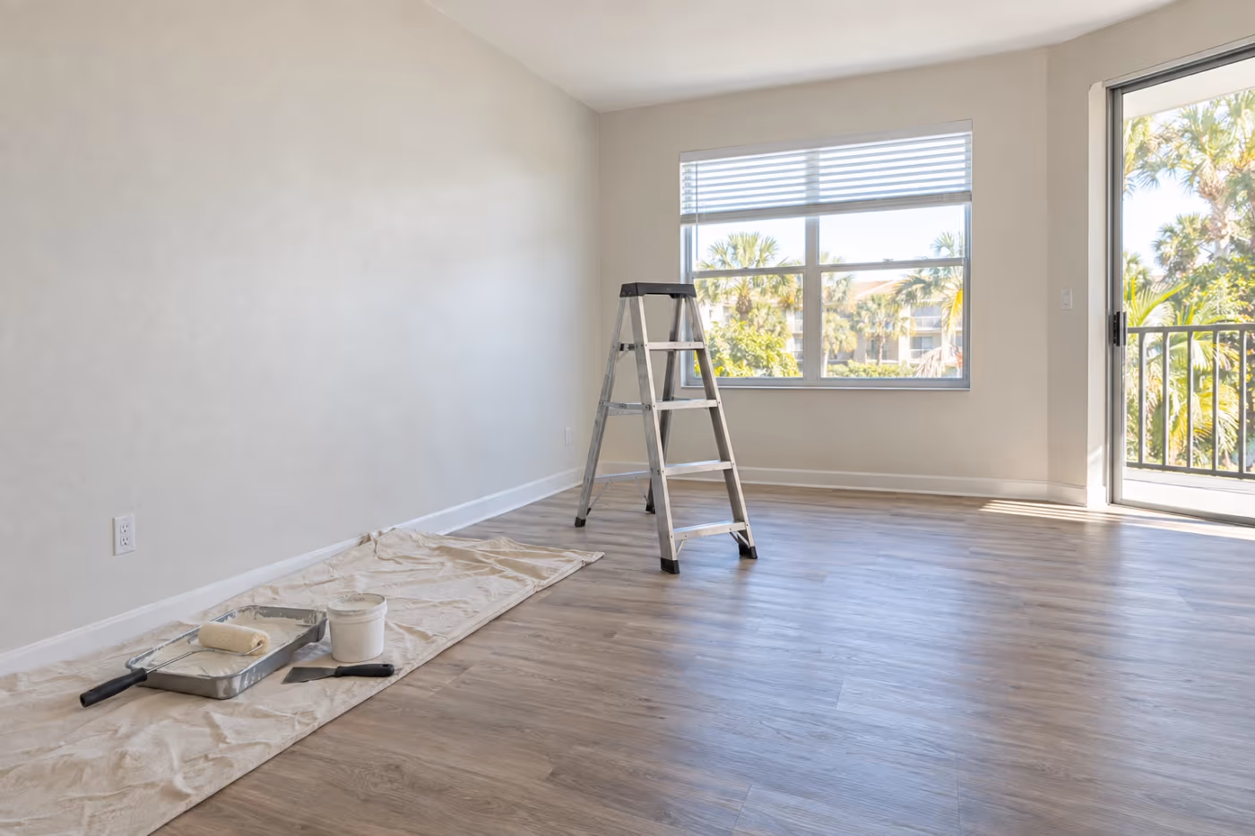 An empty Florida rental living room mid make-ready, freshly painted walls, a drop cloth with a roller tray, spackle, and a putty knife along one wall, and a step ladder standing on the plank floor