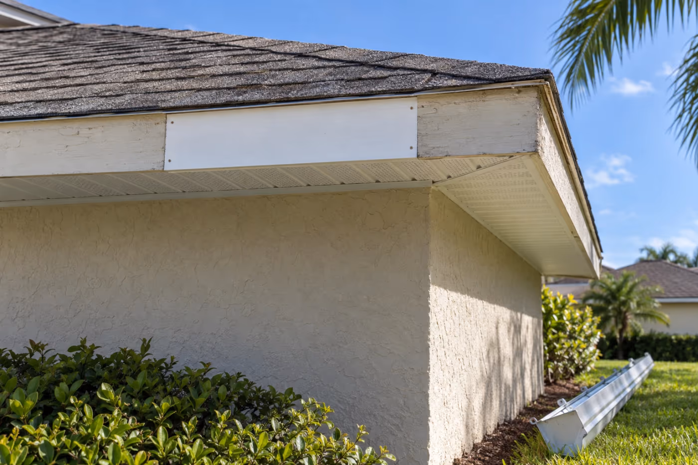 Roof edge of a Florida stucco home where a new primed white fascia board has been fastened in between older painted boards, the gutter taken down and resting on the lawn