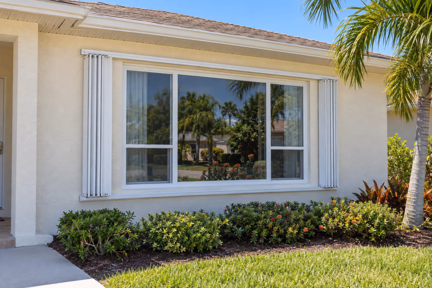 Front of a single-story Florida stucco home on a sunny day with white accordion hurricane shutters folded open on each side of a large front window, with their tracks running above and below it