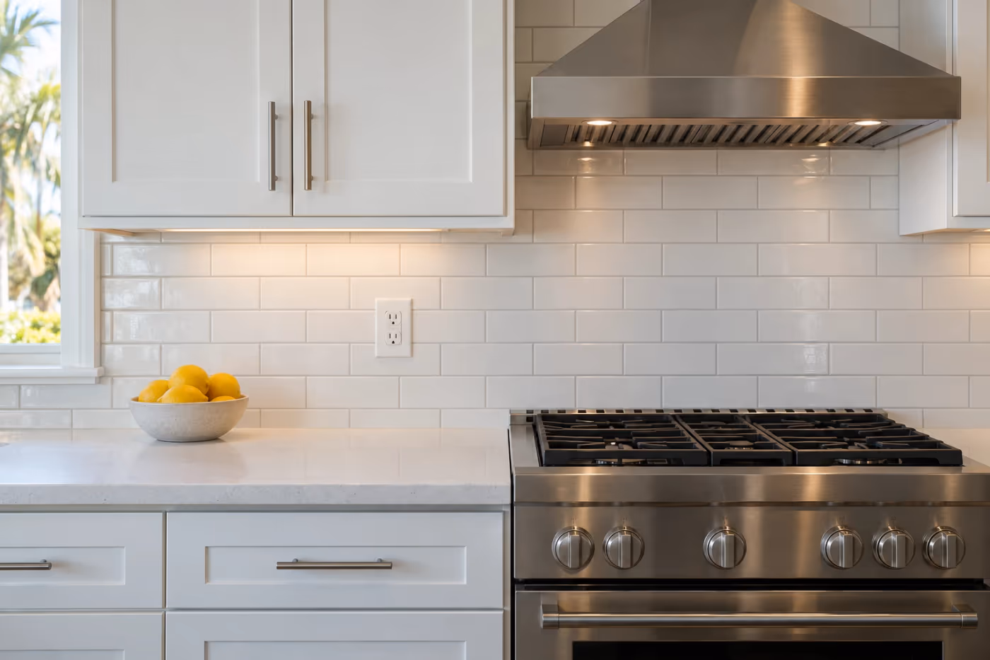 A white subway tile kitchen backsplash with light gray grout between a quartz countertop and white cabinets, cut neatly around an outlet beside a range