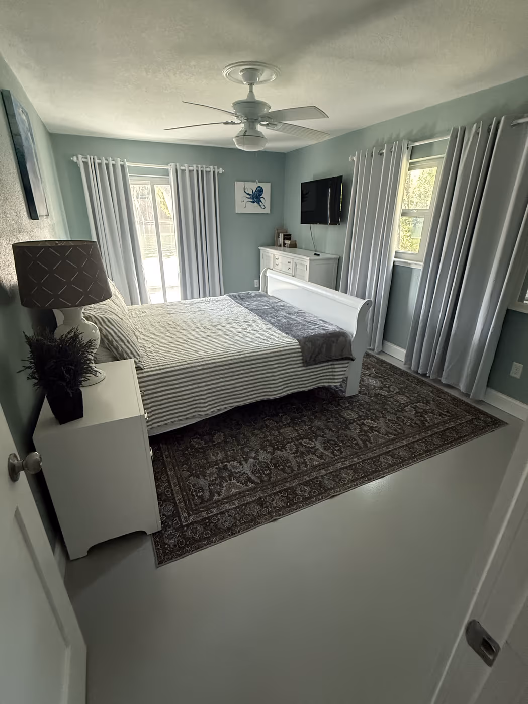 Guest bedroom with new curtain rods and gray panels on three windows, a white ceiling fan, and a wall-mounted TV
