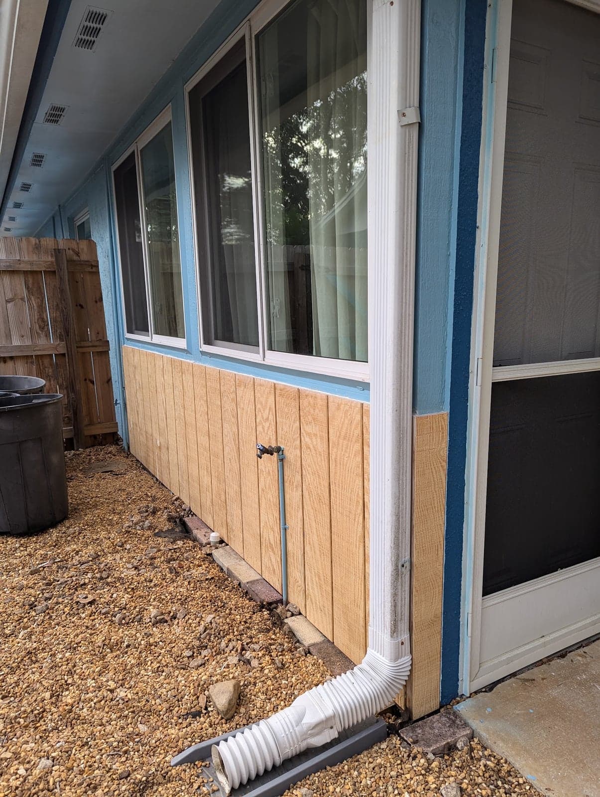 New unpainted wood siding panels installed below a row of windows on a blue house, next to a white downspout and a screen door
