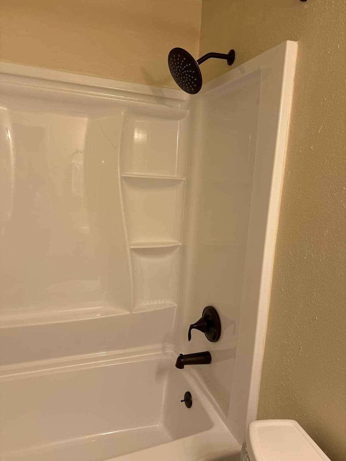 White tub and shower surround with molded shelves, an oil-rubbed bronze rain shower head, a single-lever valve, and a bronze tub spout