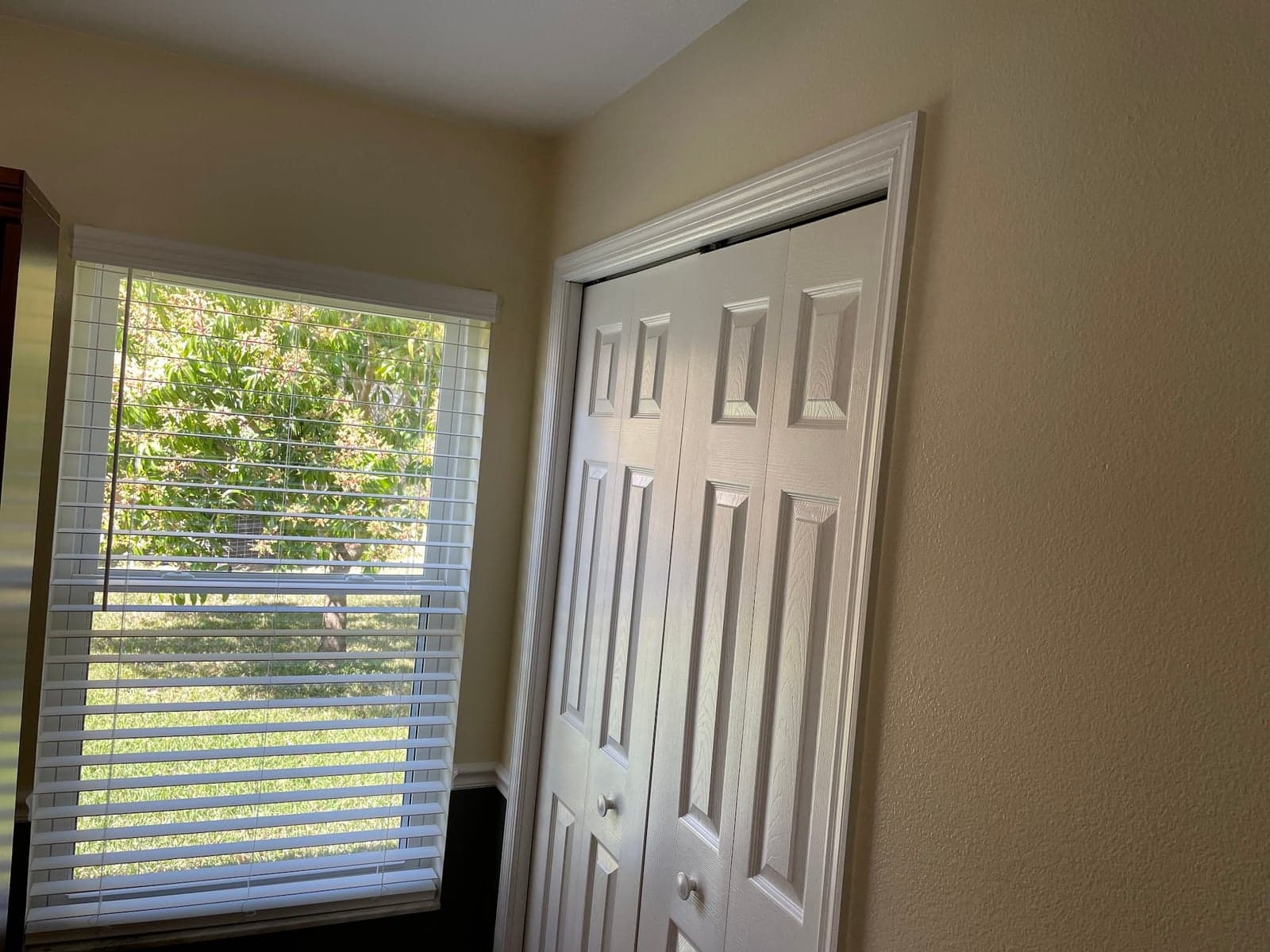 Bedroom corner with warm tan walls, white trim, white six-panel bifold closet doors, and new white wood blinds on the window