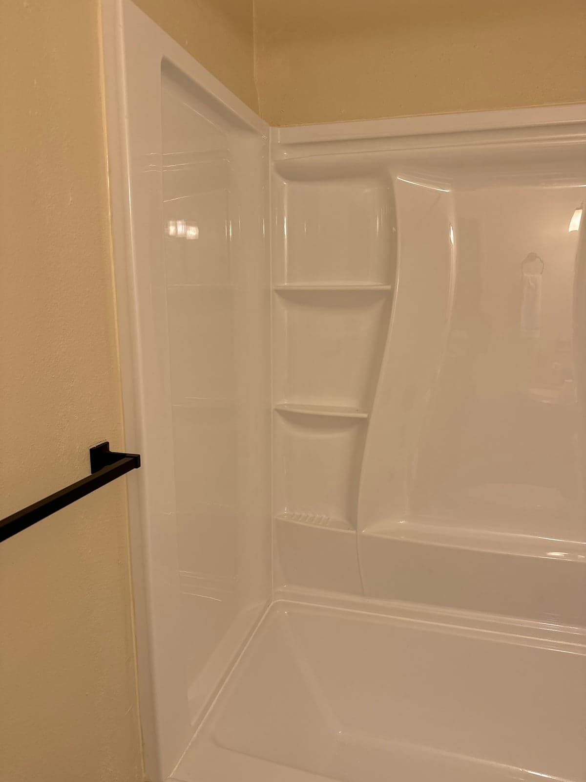 White one-piece shower surround with molded corner shelves and a curved panel, with a black towel bar on the tan wall beside it