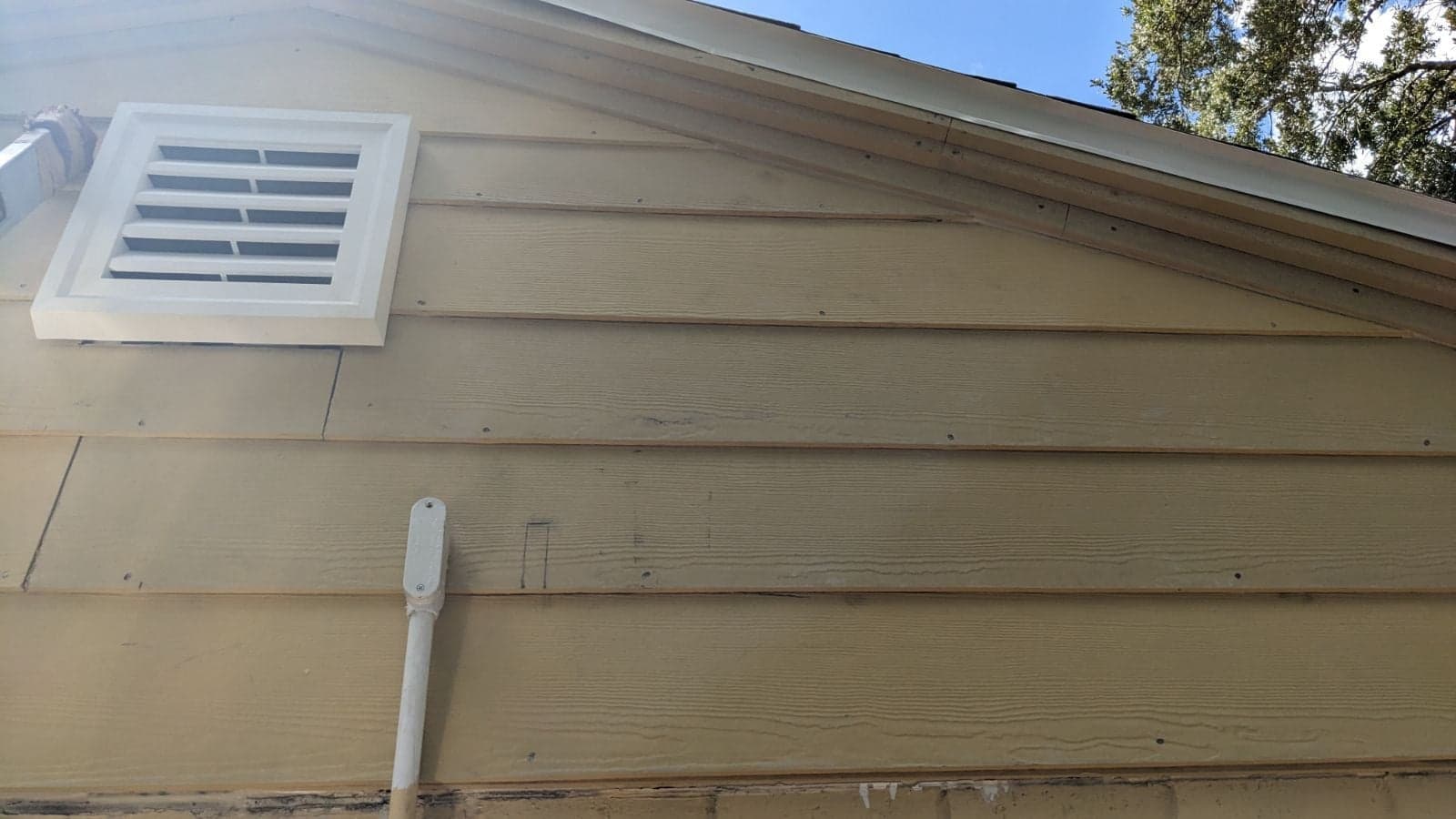 Tan lap siding on the gable end of an older home with a new white louvered vent and fresh trim boards along the roof line