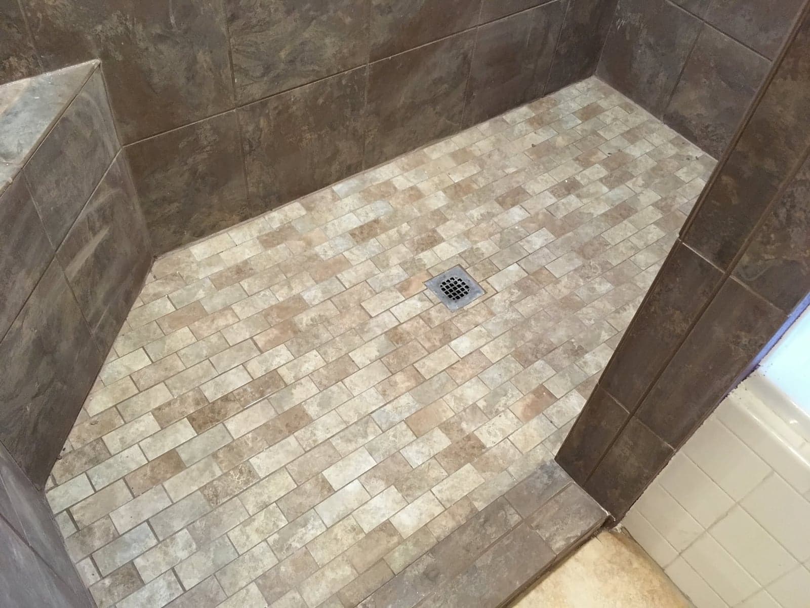 Walk-in shower with dark stone-look wall tile, a tiled bench along one side, and a beige brick-pattern mosaic floor around a square drain
