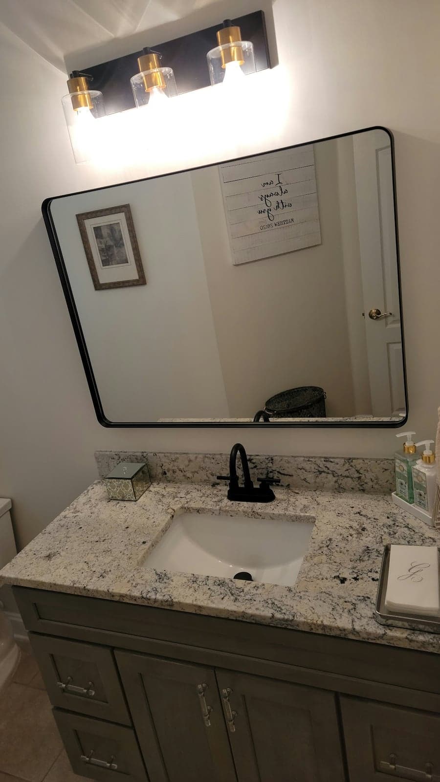 Small bathroom with a new gray vanity, granite top, black faucet, framed mirror, and a three-light vanity fixture