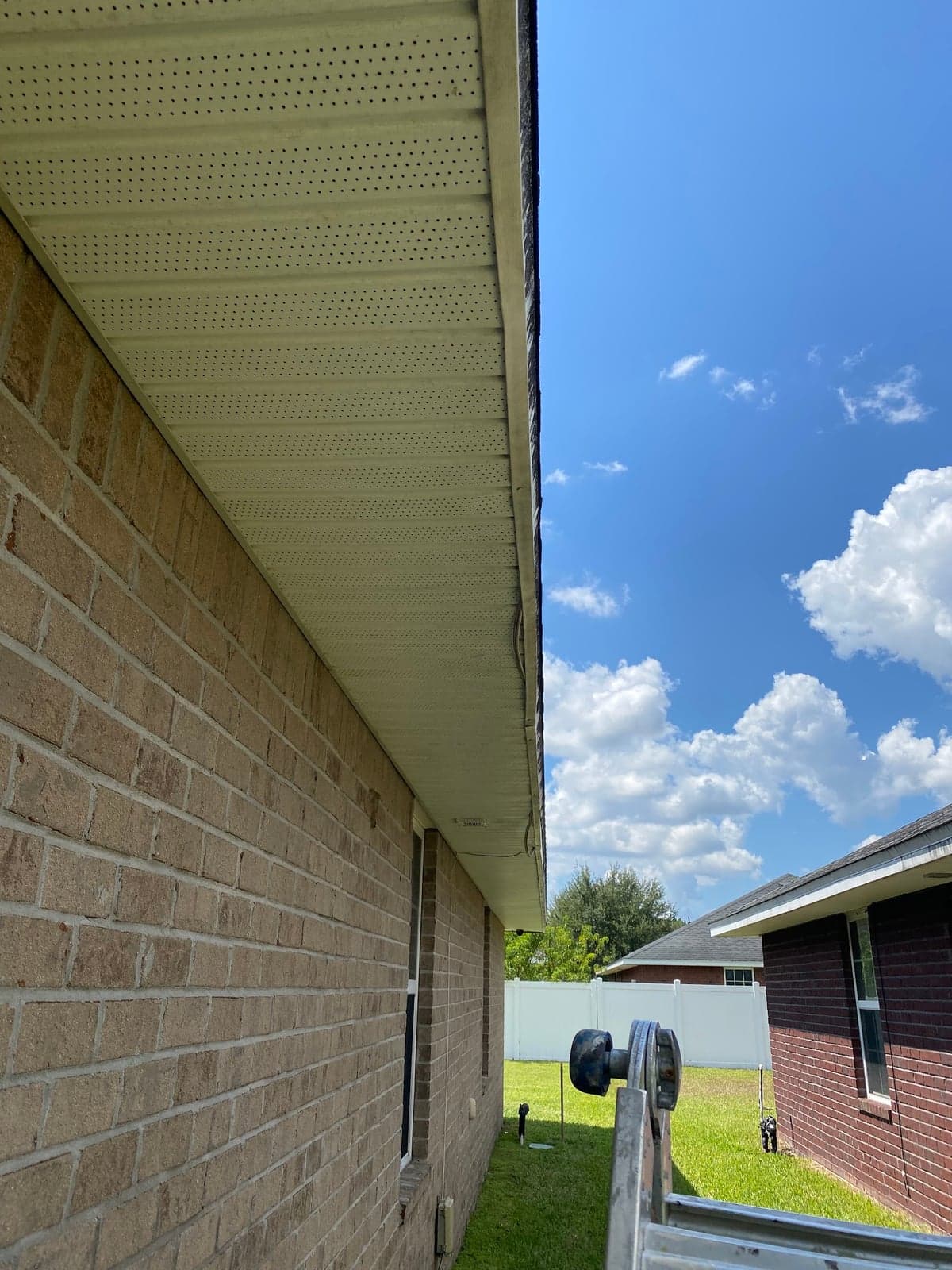 Vented white soffit and fascia running along the eave of a brick home, a ladder at the corner and a fenced yard beyond