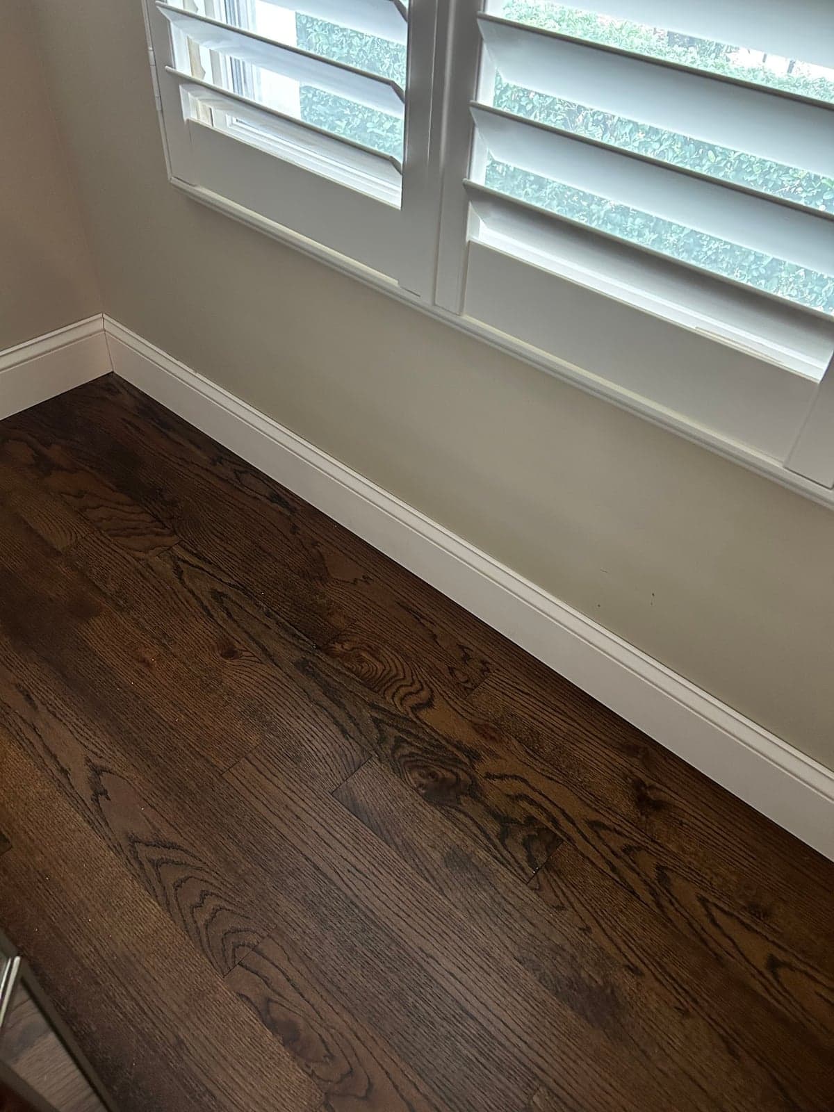Dark stained hardwood floor meeting new white baseboard under a window with white plantation shutters