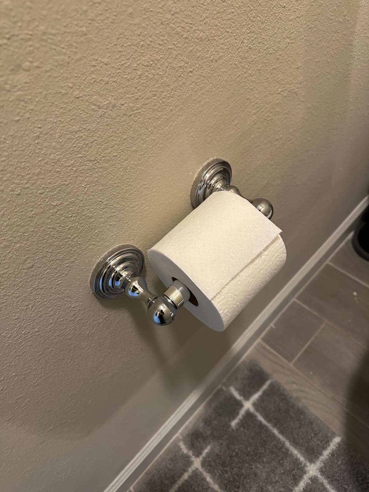 Chrome toilet paper holder mounted level on a textured beige bathroom wall above gray plank tile