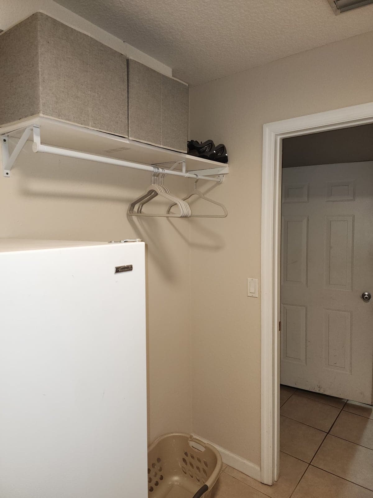 A white wire shelf with a hanging rod and clothes hangers mounted on a beige laundry room wall above a chest freezer